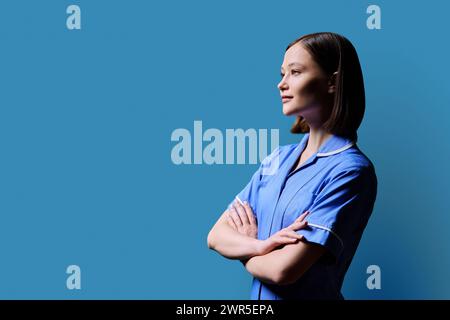 Porträt einer jungen ernsten Krankenschwester, die im Profil auf blauem Hintergrund schaut Stockfoto
