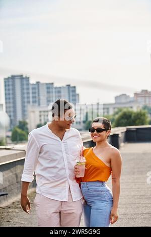 Fröhliche interrassische Freunde mit Sonnenbrille in lebendigen Gewändern, die auf dem Dach trinken und feiern Stockfoto