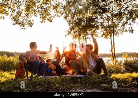 Dieses Bild strahlt die Freude einer gemischten Gruppe von Personen aus, die während der goldenen Stunde ein Picknick am See genießen. Die Gruppe mit verschiedenen Altersgruppen und ethnischen Hintergründen, einschließlich kaukasischer, schwarzer und hispanischer Personen, ist in eine entspannte, festliche Atmosphäre vertieft. Erwachsene erleben fröhliche Toasts und lebhafte Gespräche, während ein kleines Kind, begleitet von einem Teddybären, der Szene ein Element der Unschuld verleiht. Die untergehende Sonne zieht sich durch die Bäume und strahlt ein warmes, bezauberndes Leuchten über die Gruppe und den ruhigen See dahinter aus, der die perfekten Outdoor-Umgebungen umschließt Stockfoto