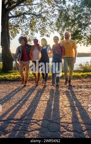 Dieses Bild fängt das entspannte Wesen einer Familie ein, die einen Spaziergang im weichen, schwindenden Licht des Abends macht. Die Gruppe ist vielfältig und umfasst Personen mit unterschiedlichem Alter und ethnischem Hintergrund, was auf eine moderne, gemischte Familie hindeutet. Von einem jungen Afroamerikaner mit lockigen Haaren bis hin zu einem älteren Mann mit weißem Bart in einem gelben Hemd ist das Gesicht jedes Menschen hell vor der Freude des Augenblicks. Sie gehen Arm in Arm, zeigen Nähe und Zuneigung. Die untergehende Sonne wirft lange Schatten vor ihnen auf den Weg, und der große Baum im Hintergrund steht als stiller Zeuge ihrer Scherbe Stockfoto