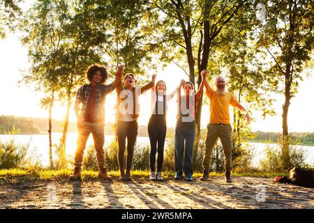 Vor dem strahlenden Hintergrund einer untergehenden Sonne verkörpert dieses Bild die temperamentvolle Feier einer Gruppe von Freunden am See. Mit triumphierend erhobenen Armen und einem hellen Lächeln über ihren Gesichtern verströmen sie einen Hauch von Sieg und purer Begeisterung. Die Gruppe ist vielfältig und umfasst Personen mit unterschiedlichem Alter, Geschlecht und ethnischem Hintergrund, einschließlich Kaukasier und Schwarzer. Das goldene Sonnenlicht umhüllt die Gruppe und erzeugt einen Silhouetteneffekt, der die festliche Stimmung unterstreicht. Der See dahinter, der von den Reflexionen des Sonnenuntergangs schimmert, verleiht dieser Momente eine ruhige und zugleich belebende Atmosphäre Stockfoto