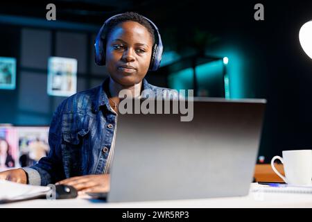 Fokussierte schwarze Frau, die schnurlose Kopfhörer mit Laptop trägt und Notizen schreibt. afroamerikanische Studentin mit Headset, die Online-Webinar ansieht und Audiokurse auf PC anhört. Stockfoto