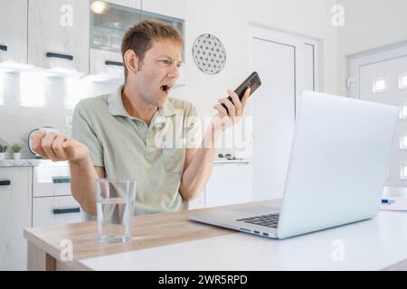 Wütender Mann streitet und schreit auf dem Handy, während er zu Hause online am Laptop arbeitet. Ein genervter Mann, der freiberuflich in die Handygeste schreit Stockfoto