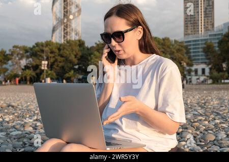 Digitale Nomadenfrau in Sonnenbrille, die mit Laptop arbeitet, sitzt am Strand am Meer während Sonnenuntergang. Freiberuflich arbeitende Frauen arbeiten aus der Ferne online Stockfoto