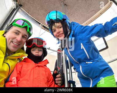 Im Inneren eines Skilifts teilen sich drei Personen, Erwachsene und zwei Kinder in Skikleidung mit Schutzbrille einen freudigen Moment beim Selfie vor der Kamera Stockfoto