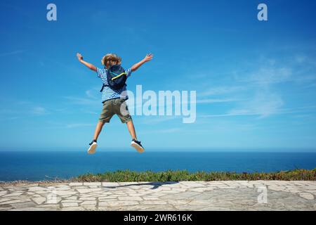 Glückliches blondes Kind mit Rucksack und Marine-T-Shirt springt hoch aufgeregt, um das Meer an heißen Sommertagen zu sehen Stockfoto