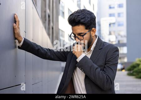 Ein junger Indianer in einem Business-Anzug steht neben dem Büro, lehnt seine Hand an die Wand und leidet an starkem Husten, bedeckt seinen Mund mit der Hand. Stockfoto