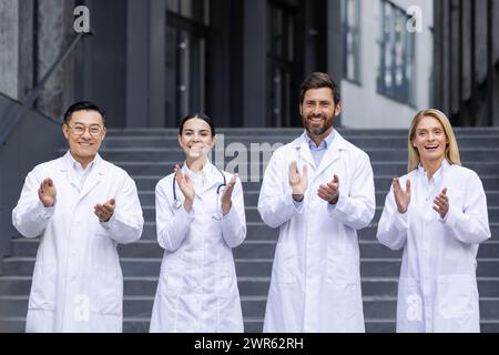 Porträt einer multirassischen Gruppe von Ärzten, die vor einer Klinik stehen, in weißen Mänteln applaudieren und lächeln, während sie in die Kamera schauen. Stockfoto