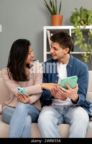 Vielfältiges junges Paar lächelt und hat Spaß mit dem Online-Gerät. Lächelnde Frau, die ein Tablet zeigt, und ein Mann, der auf einer Couch in ihrem Wohnzimmer zu Hause sitzt Stockfoto