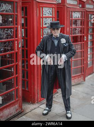 Ein stilvoller achtjähriger Mann in einem Nadelstreifenanzug posiert vor einer Reihe roter Telefondosen in London. Stockfoto