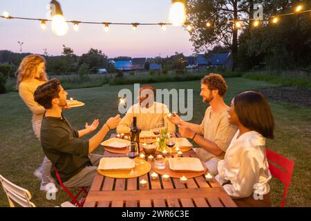 Eine intime Dinner-Party im Freien unter Freunden, bei der Sie mit Glitzern unter dem sanften Schein der Lichterketten feiern, während sich die Dämmerung über dem Garten niederschlägt. Twilight Garden Party mit Freunden und Sparklern. Hochwertige Fotos Stockfoto