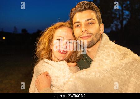 Dieses Bild strahlt Wärme und Zuneigung aus und fängt ein Paar ein, das in eine flauschige Decke gehüllt ist, ein Lächeln in der Dämmerung des Abendhimmels aus. Herzliche Umarmung: Ein Paar genießt einen gemütlichen Abend im Freien. Hochwertige Fotos Stockfoto