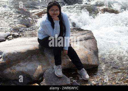 Zufriedene Frau sitzt und entspannt sich auf einem großen Felsen am sprudelnden Ganges River in Rishikesh Stockfoto