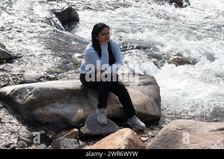 Die lächelnde Frau sitzt gemütlich auf einem übergroßen Felsen, Hand am Kinn, am sprudelnden Wasser des Ganges River in Rishikesh Stockfoto