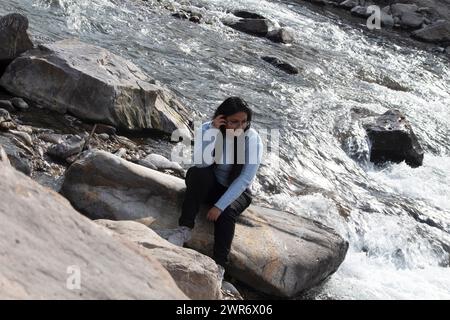 Glückliche Frau entspannt sich auf einem großen Felsen in der Nähe des Ganges in Rishikesh, lächelt und genießt die wunderschöne Landschaft Uttarakhand Stockfoto