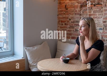 In einem ruhigen Café wird eine junge blonde Frau dargestellt, die einen Moment der Einsamkeit genießt. Sie sitzt an einem Holztisch und blickt nachdenklich aus einem großen Fenster, mit sanftem Tageslicht, das ihr Gesicht beleuchtet. Eine Tasse Kaffee wird sanft in ihren Händen gehalten, was auf eine friedliche Pause in ihrem Tag hindeutet. Die freiliegende Ziegelwand an ihrer Seite verleiht dem modernen Interieur einen rustikalen Charme und schafft eine gemütliche und einladende Atmosphäre. Die Einfachheit der Szene wird durch den Kontrast zwischen dem Blick auf die Stadt draußen und dem warmen, intimen Raum im Inneren bereichert. Besinnliche junge Frau genießt Kaffee in einem schicken Café. Hohe Qualität Stockfoto