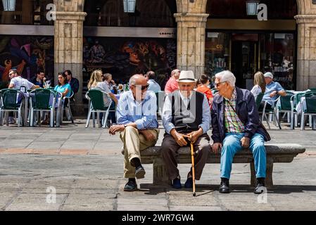 Salamanca, Spanien, 10. Juni 2023: Drei Senioren saßen auf einer Bank mitten auf der Plaza Mayor, genießen einen heißen Sommertag und unterhalten sich glücklich. Stockfoto