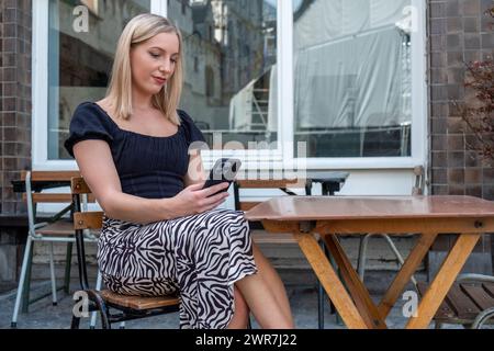Auf diesem Bild sehen wir eine ruhige junge Frau mit blonden Haaren, die in einem Straßencafé sitzt und tief mit ihrem Smartphone beschäftigt ist. Sie trägt eine modische schwarze Bluse in Kombination mit einem Rock mit Zebrastreifen und verkörpert einen lässigen, aber trendigen Look, der für die Stadt geeignet ist. Der Holztisch und die Stühle bieten eine entspannte Atmosphäre im Freien, während das Fenster des Cafés die städtische Umgebung um sie herum reflektiert. Diese Szene fängt einen privaten Moment in der Öffentlichkeit ein und zeigt die Schnittstelle zwischen persönlichem digitalem Engagement und dem geschäftigen Stadtleben. Stilvolle Frau, die ihr Smartphone im Sidewalk Cafe überprüft. Hohe Qualität Stockfoto