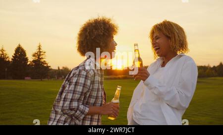 Fröhliche, multiethnische afrikanische Freundinnen mit Afro-Haaren, die Spaß haben und im Sommer bei goldenem Sonnenuntergang im Freien ein paar Biere trinken Stockfoto