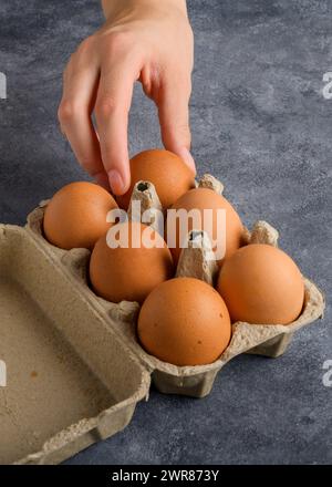 Die Hand einer Frau nimmt ein Ei aus der Eierbox, die sich auf dem grauen Küchentisch befindet, Nahaufnahme, Lebensstil, Kochblog Stockfoto