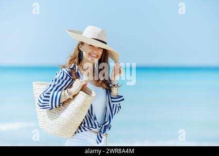 Lächelnde elegante 40-jährige Frau am Meer mit Strohsack und Strohhut. Stockfoto