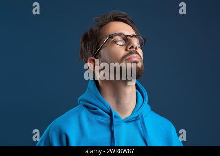 Ein heiterer junger Mann mit Brille, der seine Augen in friedlicher Haltung schließt Stockfoto