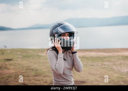 Eine Motorradfahrerin schnallt ihren Helm vor einer ruhigen Seebaumkulisse Stockfoto