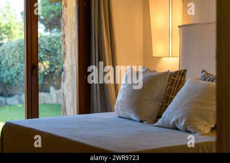 Horizontaler Blick auf ein Schlafzimmer in einer Luxusvilla in Costa Smeralda, Sardina, Italien. Riesiges Bett mit Kopfkissen, Panoramafenster mit Blick auf den Garten Stockfoto