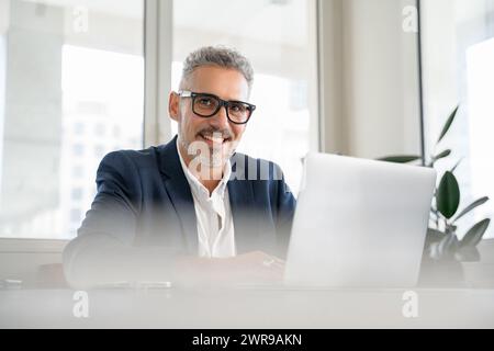 Ein selbstbewusster Geschäftsmann lächelt, während er an seinem Laptop arbeitet. Der helle Bürobereich spiegelt eine positive, energiegeladene Arbeitsatmosphäre wider. Dieses Bild zeigt die Freude an zufriedenstellender Arbeit. Stockfoto