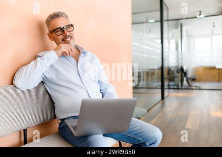 Mit einem Lächeln und einem besinnlichen Look arbeitet ein Geschäftsmann in einer entspannten Atmosphäre an seinem Laptop, die die Mischung aus Komfort und Unternehmenssinn verkörpert Stockfoto