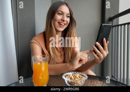 Das lachende brasilianische Mädchen sendet Nachrichten mit der Smartphone-Chat-App auf dem Balkon zu Hause. Zeit zum Entspannen. Freizeit. Stockfoto