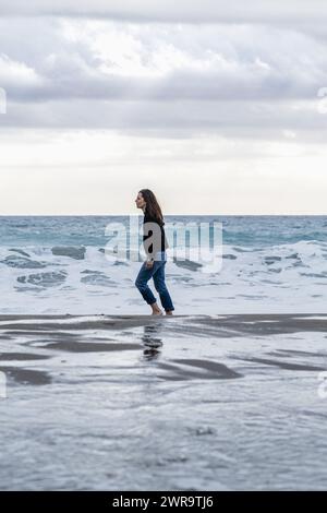 Erwachsene Frau mit langen braunen Haaren geht alleine und sorglos barfuß an der Küste. Lässige Freizeitkleidung mit durchgehender Seitenansicht zu Jeans und Winterpullover. Stockfoto