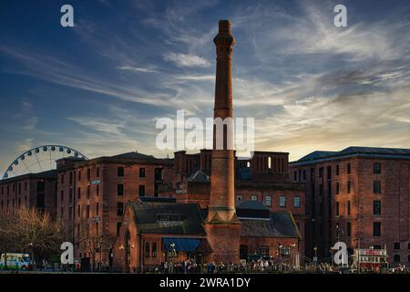 Historische Gebäude aus roten Backsteinen mit hohem Kamin vor einem dramatischen Sonnenuntergang, mit einem Riesenrad im Hintergrund in Liverpool, Großbritannien. Stockfoto