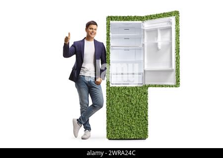 Porträt eines jungen Mannes in voller Länge, der Daumen nach oben bewegt und sich auf einen nachhaltigen Kühlschrank gestützt, isoliert auf weißem Hintergrund Stockfoto