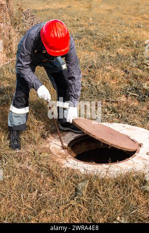 Ein Arbeiter mit einem Schutzhelm hebt einen Mannlochdeckel auf einen septischen Brunnen. Inspektion und Instandhaltung von Kanalisationssystemen im ländlichen Raum. Stockfoto