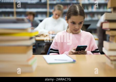 Schulmädchen benutzt Handy in der Schulbibliothek Stockfoto