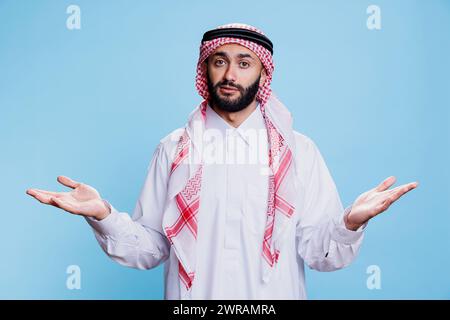 Nachdenklicher Mann mit muslimischem Dschungel und Kopftuch, stehend und zuckte Schultern, während er in die Kamera schaut. Arabische Person, die zögerlich mit offenen Armen Gesten-Studio-Porträt zeigt Stockfoto