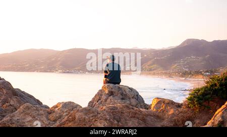 Ein junger Mann, der auf einem Felsen sitzt und den Ozean bei Sonnenuntergang überblickt. Kontemplationskonzept, reflektierend oder denkend, ruhige Meditation am Strand oder an der Küste. Stockfoto