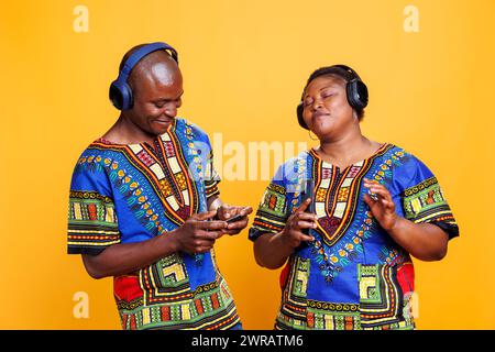 afroamerikanisches Paar genießt gemeinsam Musik über Kopfhörer und Smartphone. Frau, die Playlist mit geschlossenen Augen und zufriedenem Ausdruck hört, während der Mann auf dem Mobiltelefon den Song anschaltet Stockfoto