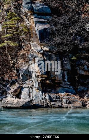 Sonnenlicht wirft Schatten auf eine felsige Klippe über einem gefrorenen Fluss, der von Bäumen gesäumt ist, in Südkorea Stockfoto