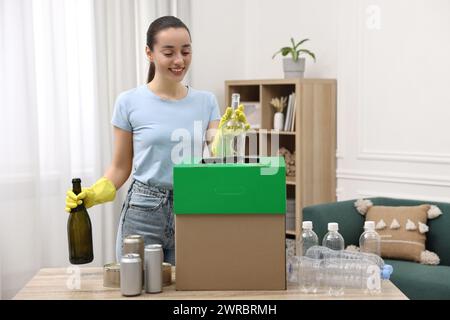 Müllsortierung. Lächelnde Frau, die Glasflasche in den Karton im Zimmer wirft Stockfoto