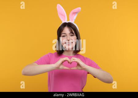 Glückliche Frau mit Hasenohren, die Herzgeste mit Händen auf orangem Hintergrund zeigt Stockfoto