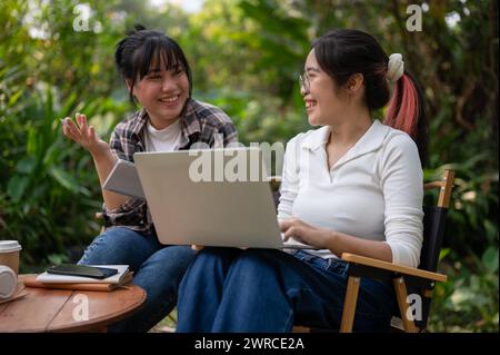 Zwei glückliche asiatische Studentinnen diskutieren über die Arbeit und machen gemeinsam Hausaufgaben auf einem Laptop, während sie an einem Tisch im Hinterhof sitzen. Stockfoto