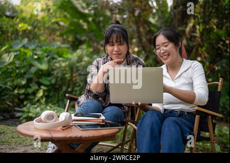 Zwei glückliche asiatische Studentinnen diskutieren über die Arbeit und machen gemeinsam Hausaufgaben auf einem Laptop, während sie an einem Tisch im Hinterhof sitzen. Stockfoto