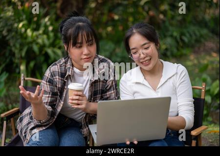 Zwei glückliche asiatische Studentinnen diskutieren über die Arbeit und machen gemeinsam Hausaufgaben auf einem Laptop, während sie an einem Tisch im Hinterhof sitzen. Stockfoto