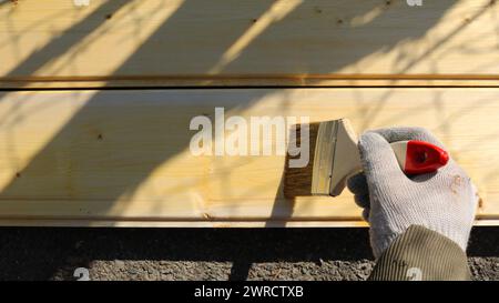 Eine Hand in einem Stoffhandschuh hält einen Pinsel und bewegt eine Schicht hellbrauner Farbe über die Oberfläche einer Platte aus frischem Holz, um sie zu imprägnieren Stockfoto