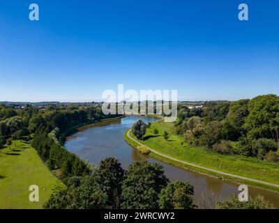 Dieses Bild bietet eine herrliche Luftaufnahme eines sich schlängelnden Flusses, der sich durch eine üppige grüne Landschaft unter dem klaren blauen Himmel schneidet. Die Flussufer sind von grünen Bäumen und Gras gesäumt, die die natürliche Kurve des Wasserwegs betonen. In der Ferne setzt sich der sanfte Fluss in Richtung Horizont fort, was auf eine kontinuierliche Fahrt von Wasser durch das Gelände hindeutet. Dieses Foto verkörpert die Harmonie von Wasser und Land aus einer erhöhten Perspektive und zeigt die Ruhe der natürlichen Welt. Panoramablick auf den River Bend. Hochwertige Fotos Stockfoto