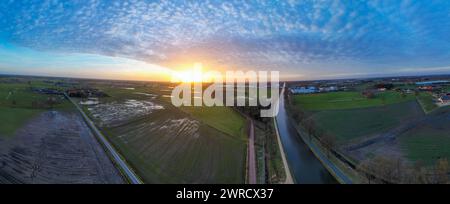 Dieses Panorama-Luftbild fängt einen atemberaubenden Sonnenuntergang mit einem Weitwinkelblick über eine ländliche Landschaft ein, die durch einen ruhigen Kanal getrennt ist. Die untergehende Sonne taucht die Szene in ein sanftes goldenes Licht und kontrastiert mit den kühlen Tönen der schattigen Felder. Der weitläufige Himmel darüber ist mit einer Wolkenstreuung geschmückt, die das letzte Tageslicht einfängt. Wohn- und Bauernhäuser sind spärlich verstreut, was zur idyllischen Landatmosphäre beiträgt. Panorama Sonnenuntergang über Kanal und ländliche Landschaft. Hochwertige Fotos Stockfoto