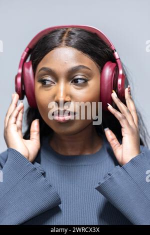 Das Bild zeigt eine junge afroamerikanische Frau, die in Musik eingetaucht ist und rote Over-Ear-Kopfhörer trägt. Ihre Augen sind sanft geschlossen und signalisieren, dass sie den Klang genießt, der ein Lieblingslied, ein fesselnder Podcast oder vielleicht eine Meditations-App sein könnte. Sie trägt ein lässiges graues Oberteil und ihr langes, dunkles Haar umrahmt ihr besinnliches Gesicht. Der neutrale Hintergrund sorgt dafür, dass der Schwerpunkt auf ihrem ruhigen Ausdruck und den lebendigen Kopfhörern bleibt. Junge Schwarze Frau, die Musik mit Kopfhörern genießt. Hochwertige Fotos Stockfoto