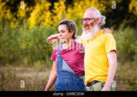 Dieses Bild zeigt einen kaukasischen Mann und eine Latina-Frau, die einen friedlichen Moment in der Natur genießen. Der Mann mit langem weißem Bart und Brille steht hinter der Frau, die ein Kleid aus Denim und ein rosa Hemd trägt, und blickt mit nachdenklichem Ausdruck in die Ferne. Er hat seinen Arm um sie herum in einer unterstützenden Geste, und sie stützt ihren Arm auf seinem, was auf eine enge und bequeme Beziehung hinweist. Die Kulisse aus üppigem Grün, das in das sanfte Licht der untergehenden Sonne getaucht ist, trägt zur reflektierenden und ruhigen Stimmung der Szene bei. Besinnliches Paar, das einen Spaziergang bei Sonnenuntergang genießt. Hohe Qualität p Stockfoto